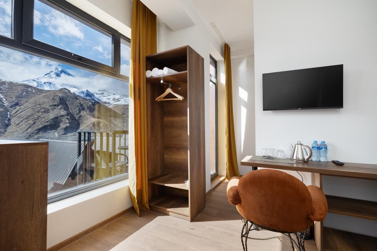 Una habitación moderna con un armario de madera, una silla marrón, un televisor de pantalla plana y una ventana con vista a la montaña.