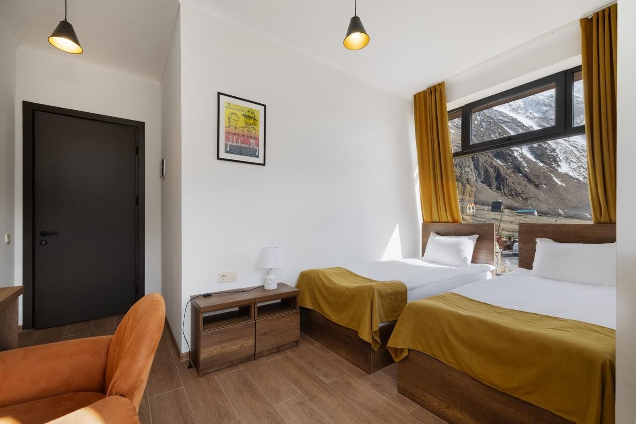 A hotel room with two beds, a desk, a chair, a window with a mountain view, and a door.