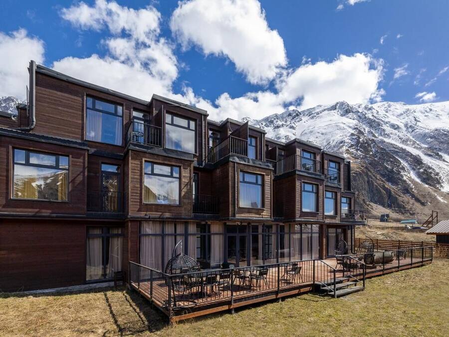 Un edificio moderno de varios pisos con amplias ventanas, un balcón y vistas a montañas nevadas.