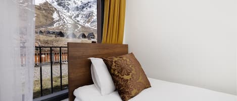 A bed with white bedding and a patterned pillow, a wooden headboard, and a window with a view of snow-capped mountains and buildings.