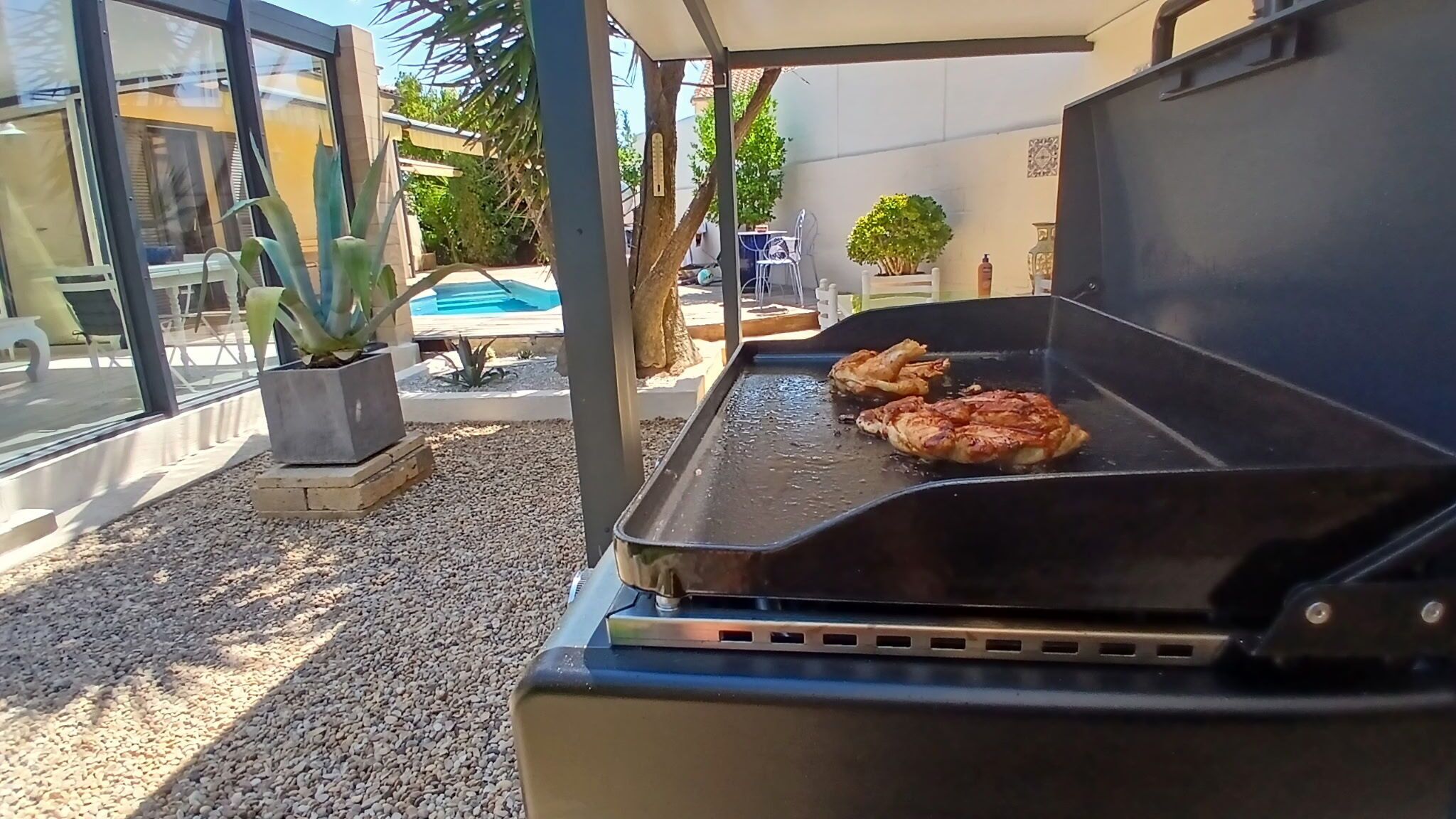 Un barbecue extérieur avec deux morceaux de viande en train de cuire, entouré d’un espace patio comprenant une piscine, des chaises et des plantes en pot.