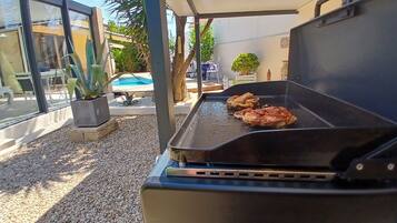 Un barbecue extérieur avec deux morceaux de viande en train de cuire, entouré d’un espace patio comprenant une piscine, des chaises et des plantes en pot.