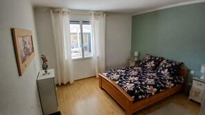 A bedroom with a wooden bed, a window with curtains, a bedside table with a lamp, and a framed picture on the wall. - VILLA DU SUD (Boujan-sur-Libron)