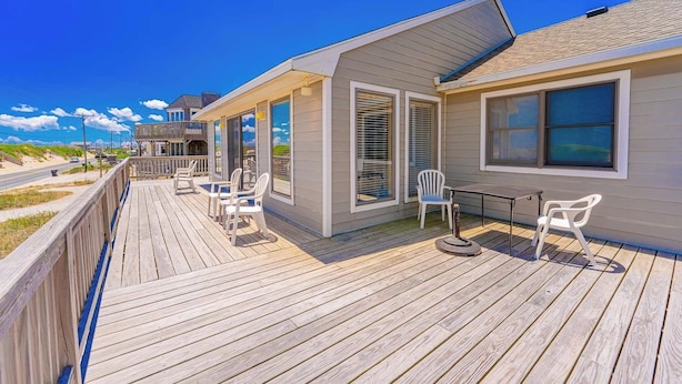 Eine Holzterrasse mit Sitzgelegenheiten im Freien, ein überdachter Bereich mit Fenstern und Blick auf den Strand.