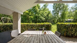 Eine Terrasse im Freien mit Holztisch und -stühlen, umgeben von Grünpflanzen und Blick auf ein Haus im Hintergrund.