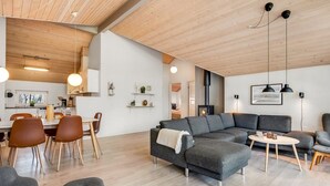 Ein modernes Wohnzimmer mit Ecksofa, Essbereich mit Stühlen, einer Küche im Hintergrund und einem Kamin.