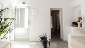 A modern, minimalist room with a white color scheme, a hanging light fixture, a door, a wall-mounted air conditioner, a potted plant, and a view into another room. - Santorinico villas  (Imerovigli)