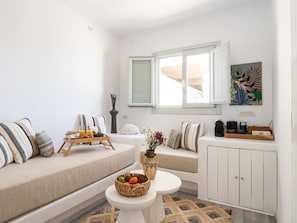 A modern living room with a sofa, a small table with a fruit bowl, a coffee station, and a window with a view.