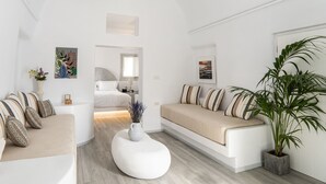 A modern, minimalist living room with a sofa, ottoman, and a large plant. - Santorinico villas  (Imerovigli)