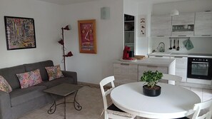 A modern living room with a sofa, a small table, a potted plant, and a kitchen area with cabinets and appliances. - Charming T2 near Avignon festival (Montfavet)