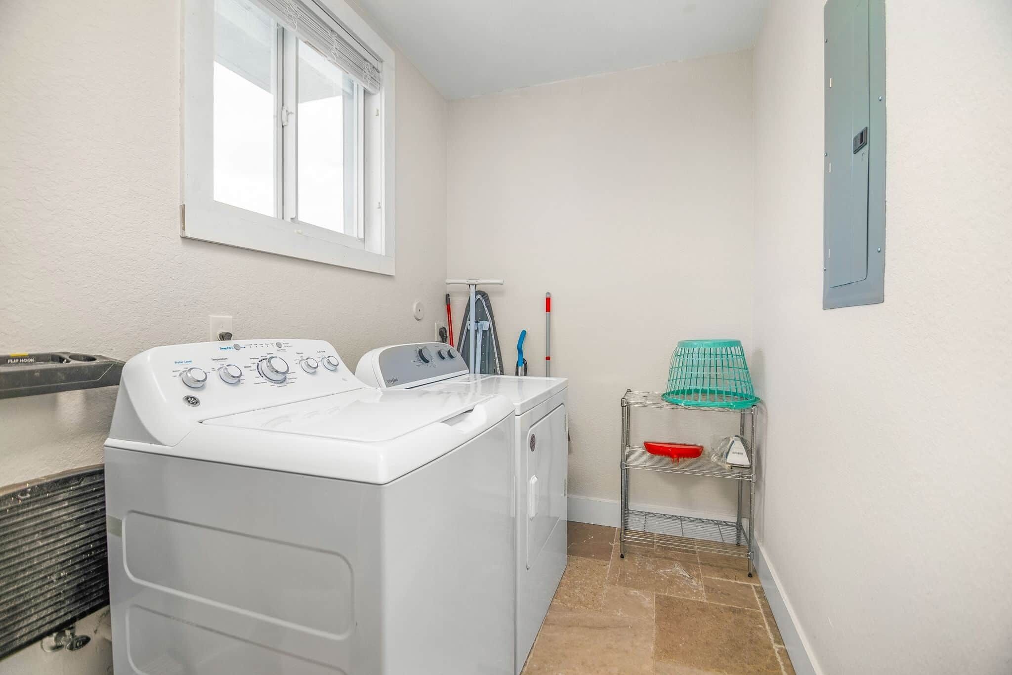 A laundry room with a front-loading washer and dryer, a window, a metal shelf with laundry baskets, and a wall-mounted electrical panel.