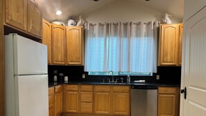 A kitchen with wooden cabinets, a white refrigerator, a dishwasher, and a window with patterned curtains. - Pacific Coast Highway Lily House (San Luis Obispo)