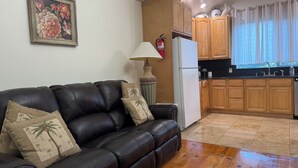 A living room with a black leather sofa, wooden cabinets, a white refrigerator, and a kitchen area with wooden countertops and a window with curtains. - Pacific Coast Highway Lily House (San Luis Obispo)