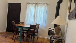 A dining area with a wooden table and chairs, a black door, a window with curtains, a wall-mounted lamp, and a fireplace. - Pacific Coast Highway Lily House (San Luis Obispo)