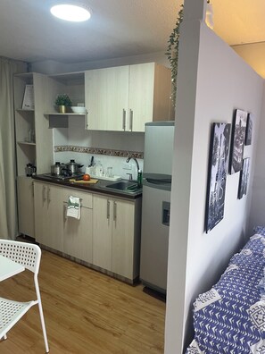 A compact kitchen with wooden cabinets, a refrigerator, and a sink. There is a dining area with a white chair and a patterned sofa. - Cozy studio apartment in the Eucharistic Quarter. (Cali)