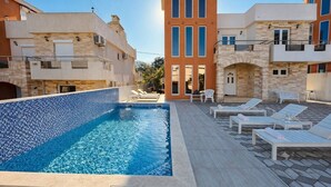 Una piscina con sillones de descanso y un deck con baldosas de piedra, rodeada de edificios de varios pisos con balcones.