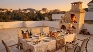 Un espacio para comer en la azotea con una mesa dispuesta para seis personas, rodeada de sillas, una chimenea de piedra y vistas a la ciudad al fondo.