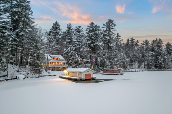 A snow-covered landscape with a house and dock, surrounded by tall trees.