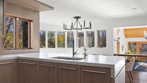 Una cocina moderna con una isla grande, una lámpara de araña y varias ventanas con vistas a follaje otoñal.