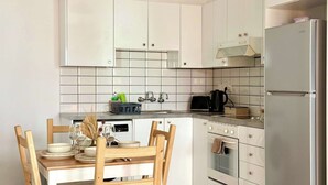 A modern kitchen with white cabinets, a stainless steel refrigerator, a dining table with chairs, and a tiled backsplash. - Coastal 2BR Apt in Mackenzie Steps from Beach (Makenzy)
