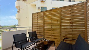 A balcony with a wooden partition, two black chairs, a small table with a plant, and an air conditioning unit. - Coastal 2BR Apt in Mackenzie Steps from Beach (Makenzy)