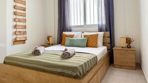 A bedroom with a bed, a bedside table, a hanging wooden rack, and a window with curtains. - Coastal 2BR Apt in Mackenzie Steps from Beach (Makenzy)