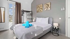 Een moderne slaapkamer met een grijs bed, witte kussens, een grijs hoofdeinde, een nachtkastje met een lamp, en een raam met gordijnen.