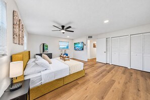 A bedroom with a bed, bedside table, lamp, ceiling fan, and a view of a beach through the window. - 6 King Suites | Pool + Game Room Oasis Near Beaches (Seminole)