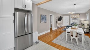 Uma cozinha moderna com eletrodomésticos de aço inoxidável, uma sala de jantar com mesa e cadeiras brancas, e uma sala de estar com sofá e televisão.