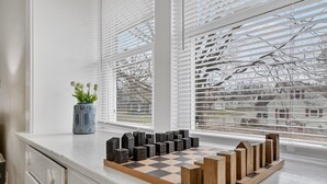 Um tabuleiro de xadrez sobre uma bancada branca, perto de uma janela com persianas, um vaso com flores, e vista para árvores e edifícios lá fora.