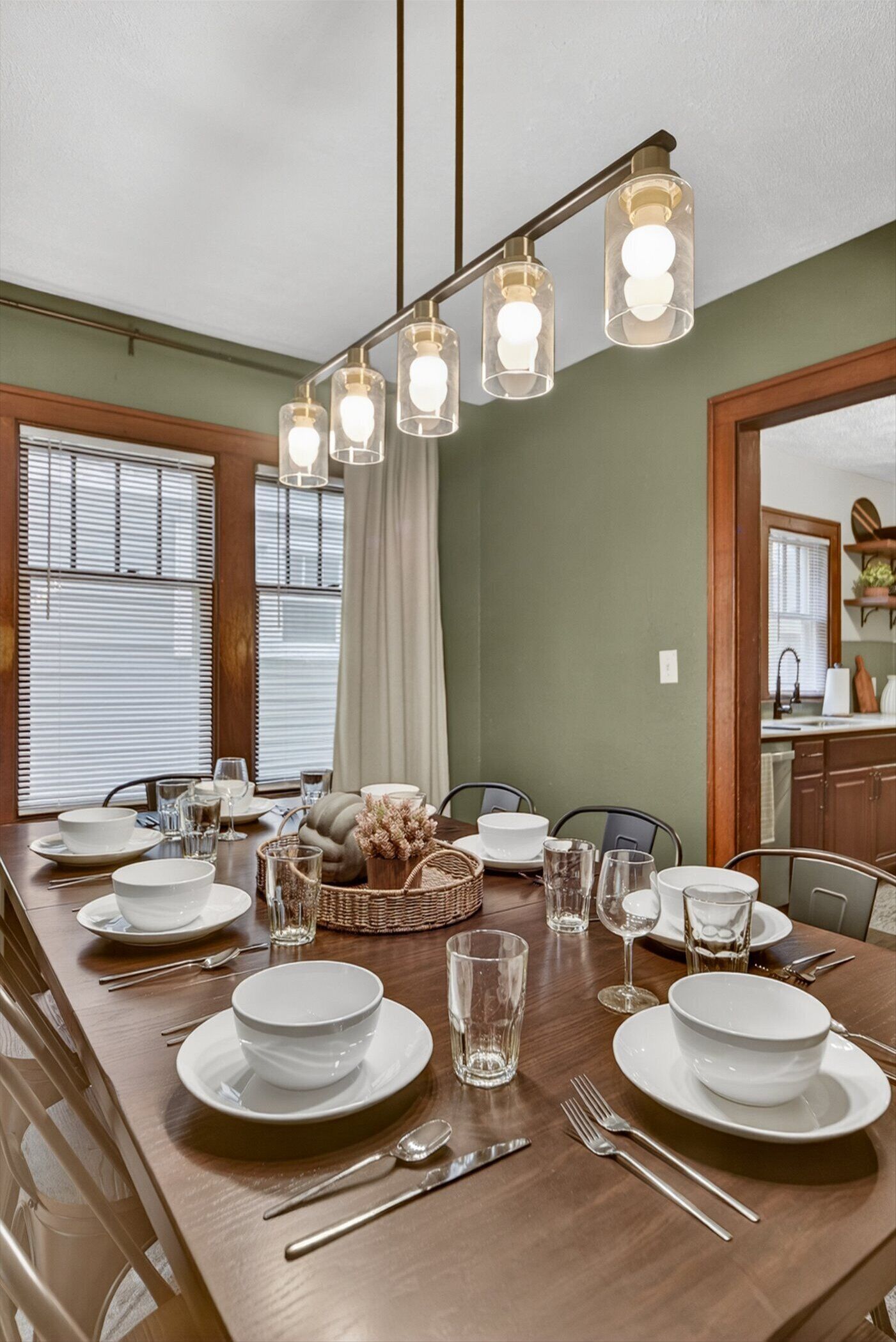 Un coin repas avec une longue table en bois, dressée pour quatre personnes, et agrémentée d’assiettes blanches, de verres et de couverts.