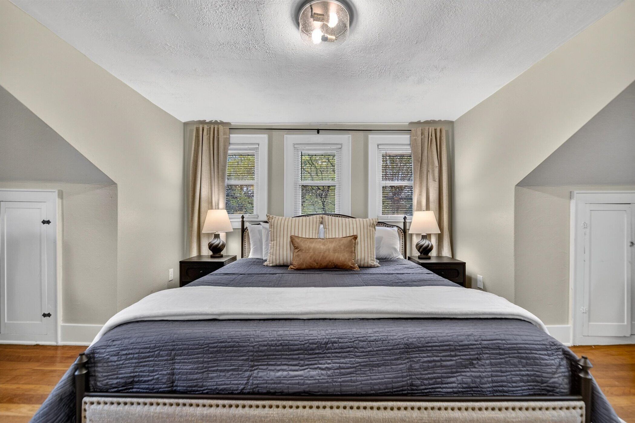 Une chambre à coucher avec un grand lit, deux tables de chevet avec des lampes, un luminaire au plafond et trois fenêtres avec des rideaux.