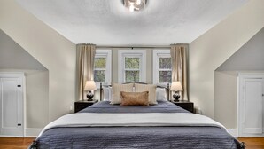 A bedroom with a large bed, two bedside tables with lamps, a ceiling light fixture, and three windows with curtains.