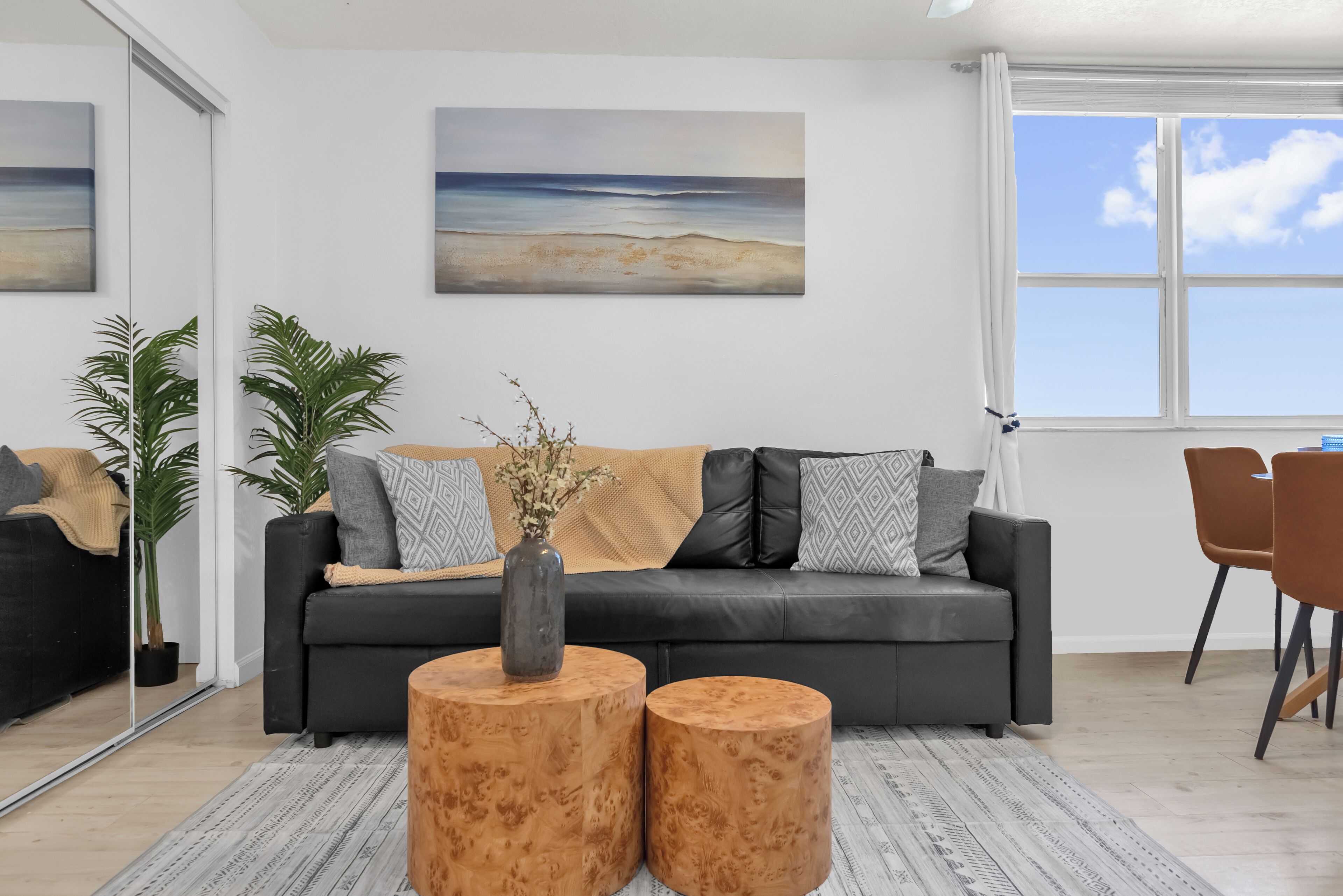 Una sala de estar moderna con un sofá negro, dos mesitas de madera, un ventanal con vistas y un cuadro en la pared.