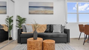 A modern living room with a black sofa, two wooden side tables, a large window with a view, and a painting on the wall. - The Vista: Oceanfront studio~View~King bed~Pool~Gym (Daytona Beach)