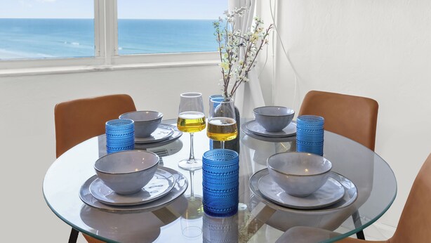 A dining table with bowls, glasses, and a vase with flowers, set against a view of the ocean. - The Vista: Oceanfront studio~View~King bed~Pool~Gym (Daytona Beach)