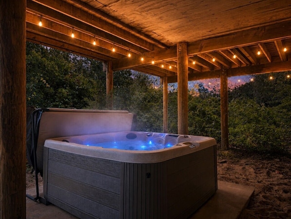 A hot tub under a wooden pergola with string lights, surrounded by greenery.