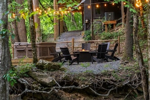 Un espace extérieur aménagé avec des sièges, un foyer et entouré d’arbres et d’une terrasse en bois.