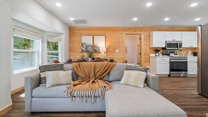 A modern living room with a grey sectional sofa, wooden panel walls, a kitchen with white cabinets and stainless steel appliances, and large windows.