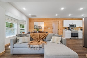 A modern living room with a grey sectional sofa, wooden panel walls, a kitchen with white cabinets and stainless steel appliances, and large windows.