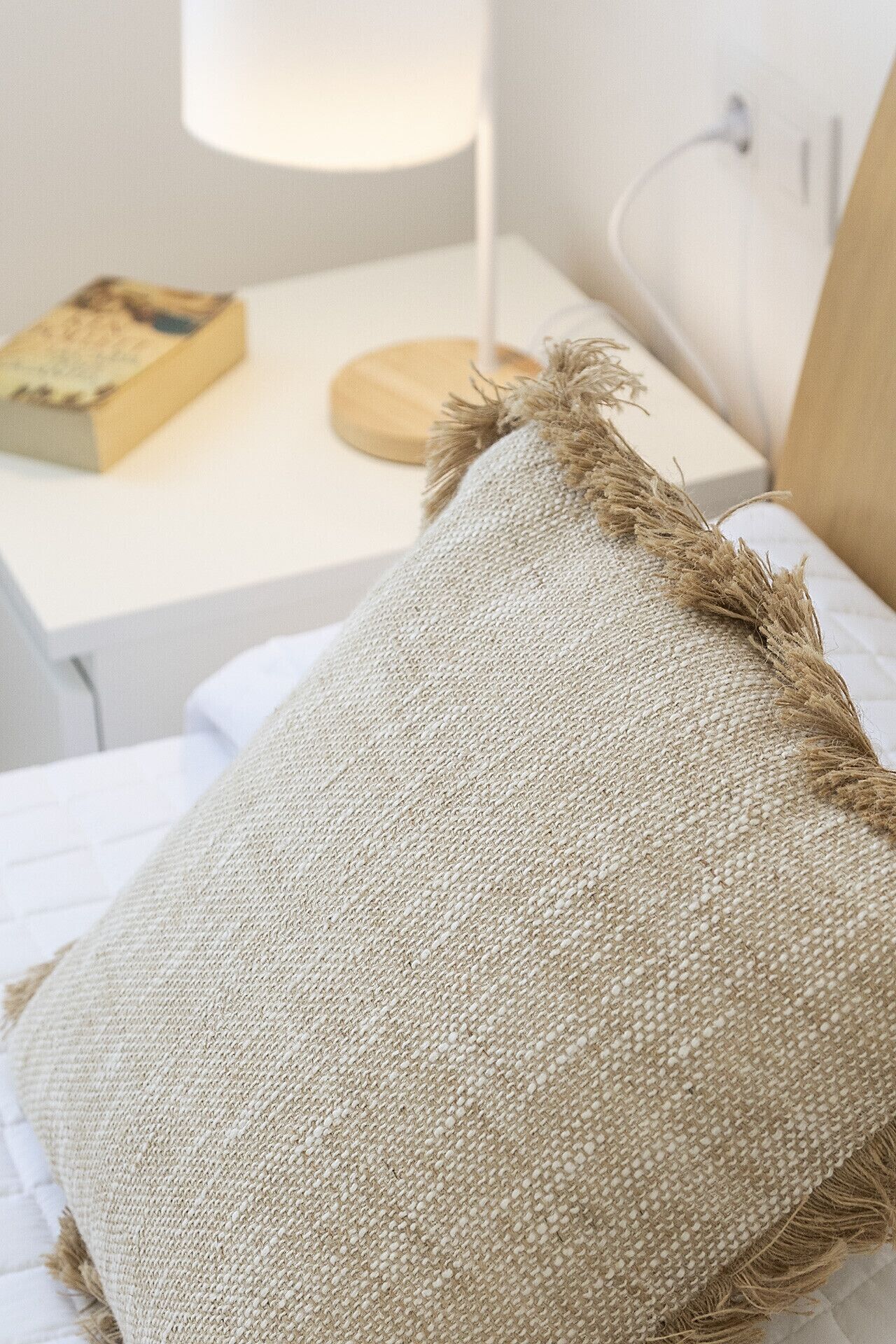 Una cama con un cojín texturizado y una mesita de noche con una lámpara y un libro.