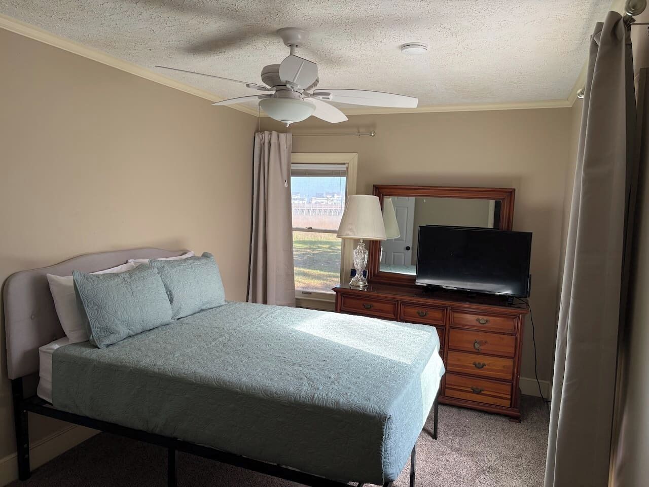Un dormitorio con una cama, un televisor en una cómoda, un ventilador de techo y una ventana con cortinas.