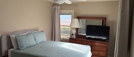 Un dormitorio con una cama, un televisor en una cómoda, un ventilador de techo y una ventana con cortinas.
