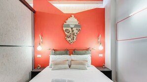 Una habitación de hotel con una cama, dos mesitas de noche, un adorno en la pared y una mesita con un florero.