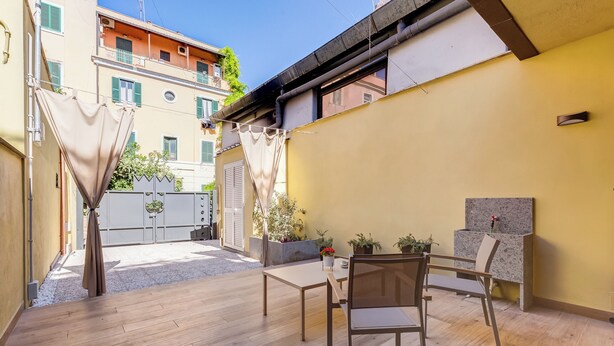 Un patio con mesa y sillas, un pequeño jardín y un edificio con balcones.