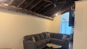 A living room with a gray sectional sofa, a glass coffee table, wooden flooring, a ceiling fan, and a TV mounted on the wall. - Superb 2-bedroom country house in charming Pittsburgh with AC (Pittsburgh)