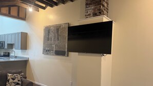 A modern living room with a flat-screen TV mounted on the wall, a sofa with a geometric-patterned pillow, and a kitchen area with grey cabinets and a microwave. - Superb 2-bedroom country house in charming Pittsburgh with AC (Pittsburgh)