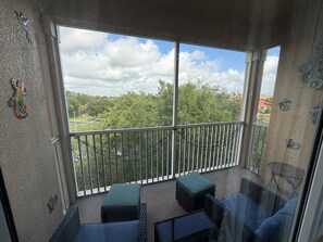 Un balcon donnant sur une route et sur un espace verdoyant, une table en verre et deux coins salon avec des coussins.