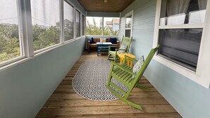 Een overdekt terras met een houten plafond, grote ramen, een rond geweven vloerkleed en groene en gele stoelen.