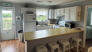 Een keuken met witte kastjes, een koelkast, een fornuis en een spoelbak. Er staan drie barkrukken bij het aanrecht.